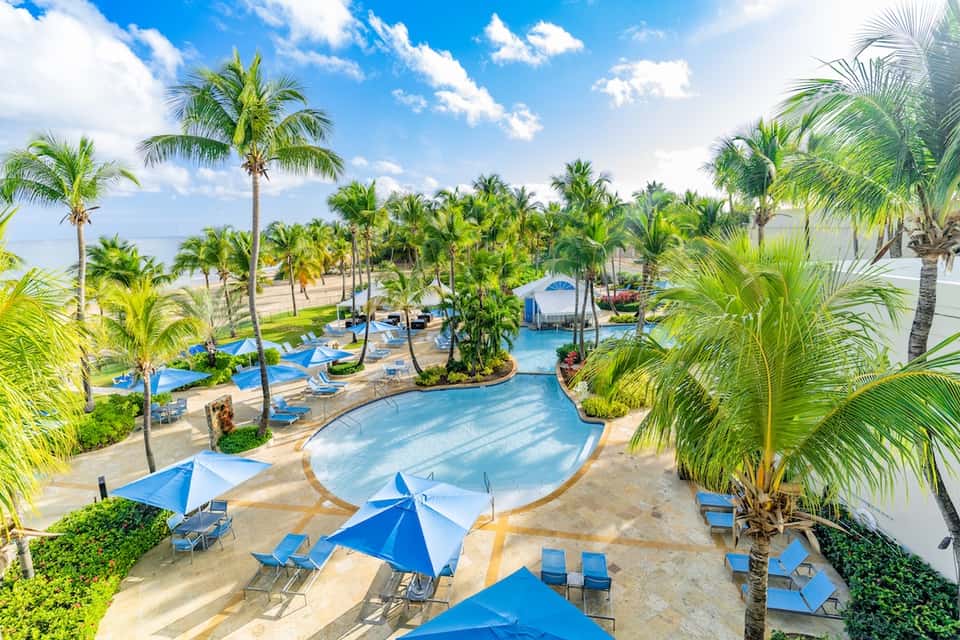 Aerial view of tropical resort with multiple pools, palm trees, blue umbrellas, and coastal setting
