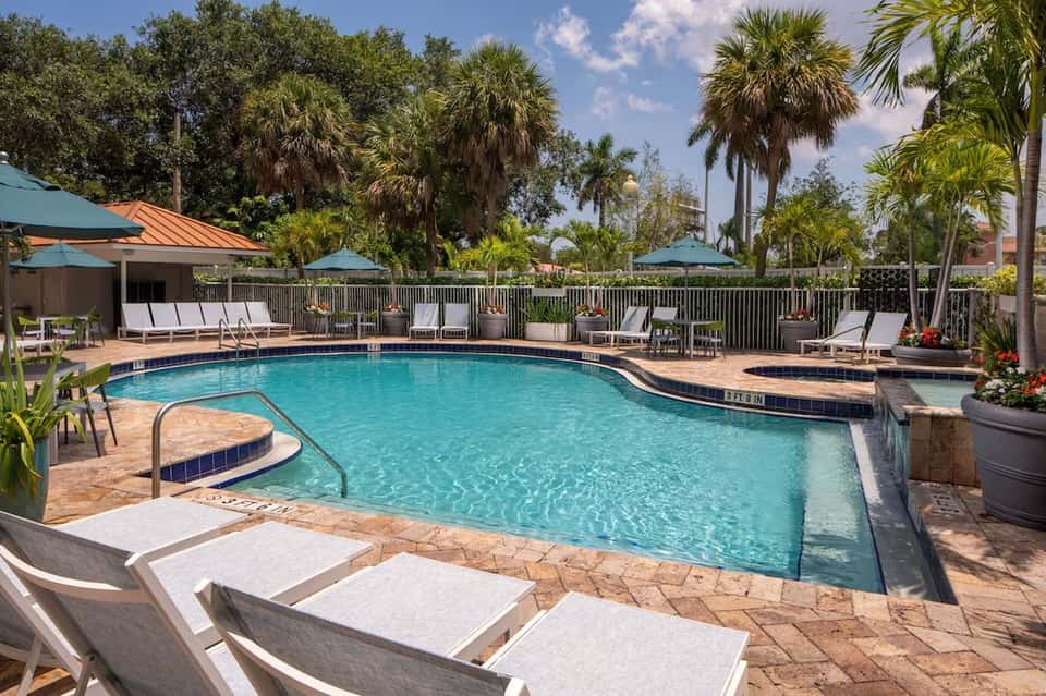 Large resort pool surrounded by lounge chairs, umbrellas, palm trees, and lush landscaping at midday