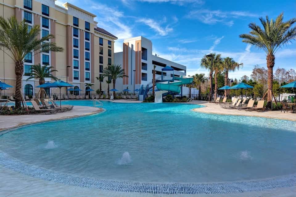 Resort swimming pool with shallow beach entry, lounge chairs, umbrellas, and modern hotel buildings