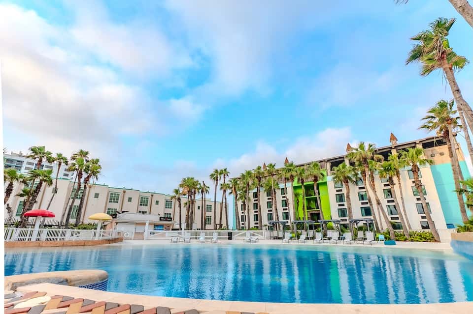 Resort pool with colorful hotel facade, palm trees, white lounge chairs, and bright sunny day at waterfront