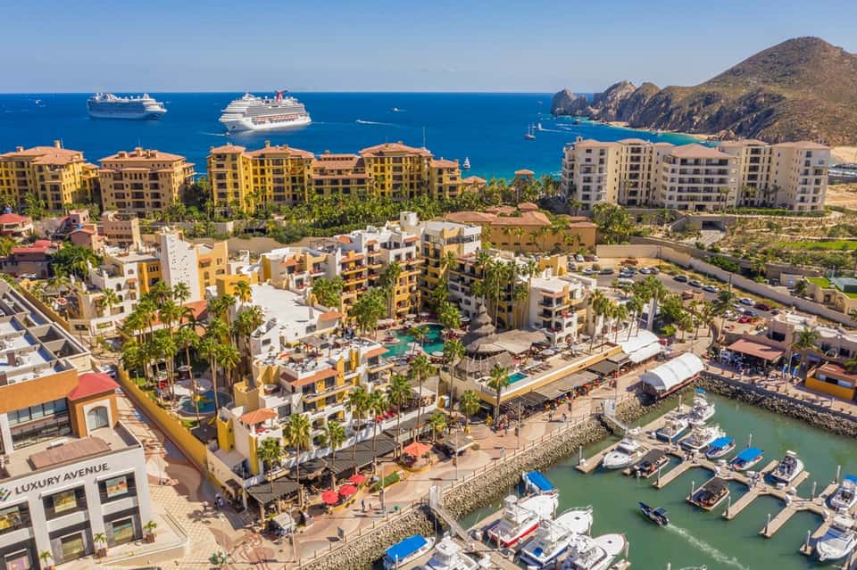 Aerial view of colorful beachfront resort with pools, palm trees, boats, and cruise ships at sea