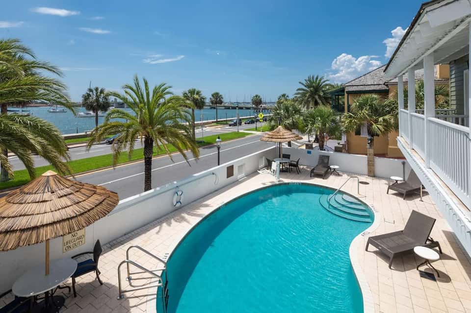 Curved swimming pool on waterfront patio with palm trees, ocean view, and white buildings