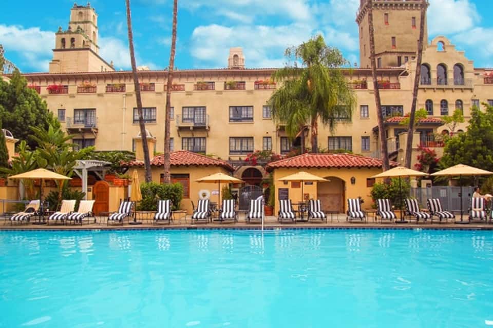 Historic Spanish-style hotel with yellow stucco facade overlooking large swimming pool with lounge chairs and umbrellas