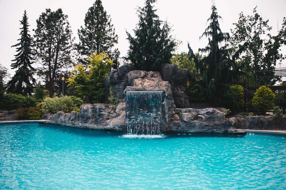 Turquoise pool with waterfall cascading from large rock formation surrounded by tall evergreen trees
