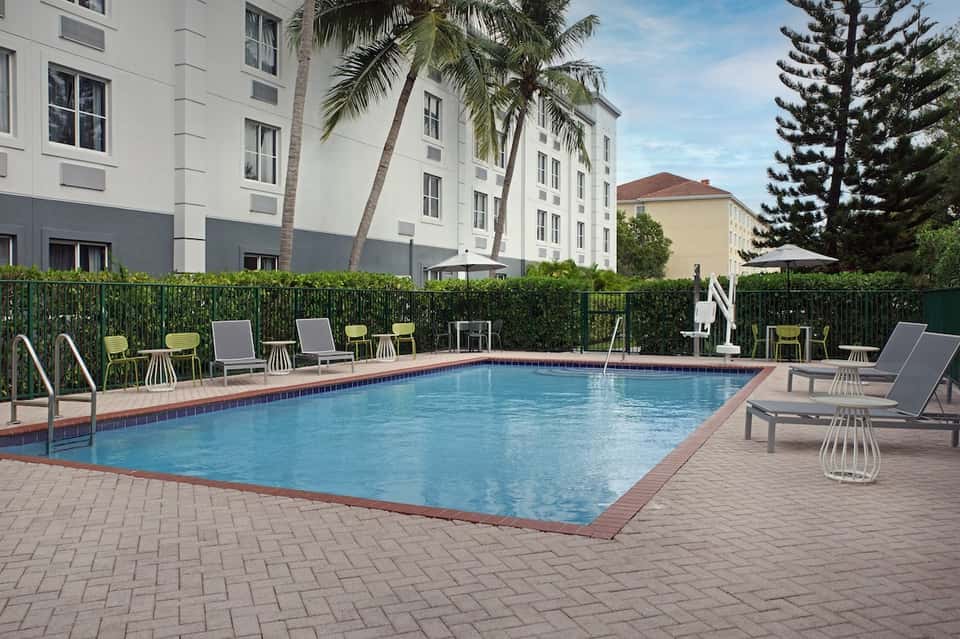 Resort rectangular pool with brick surround, loungers, high-rise hotel building backdrop, and palm trees lining fence