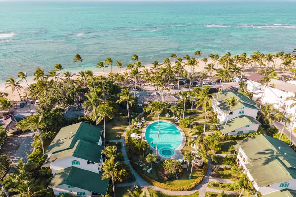 Aerial view of beachfront resort with turquoise pool, palm-lined beach, ocean, and green roofs