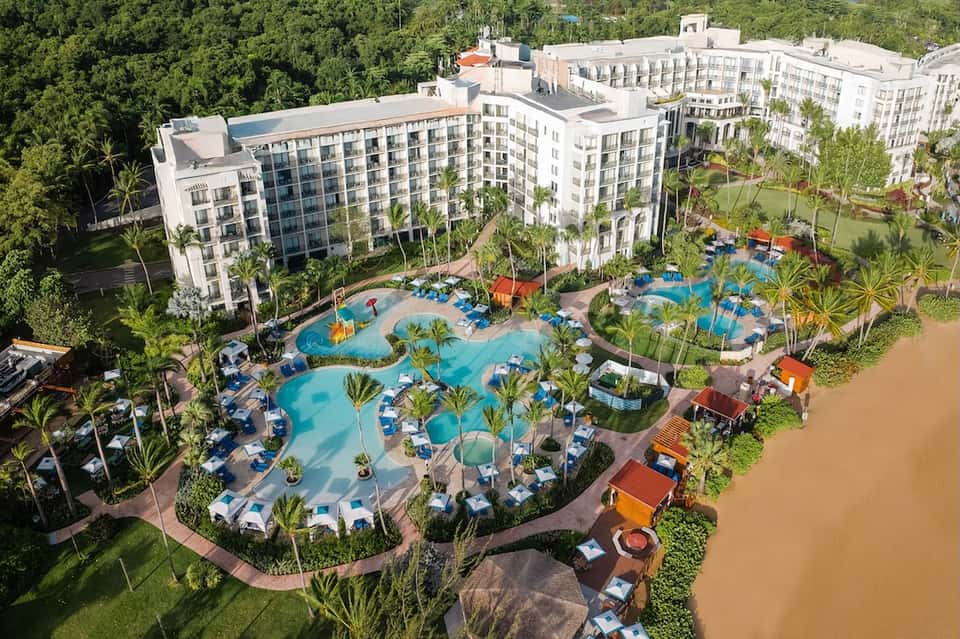 Aerial view of resort with multiple swimming pools, palm trees, lounge chairs, and modern white buildings
