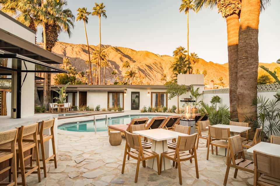 Desert resort courtyard with turquoise pool, wooden loungers, palm trees, and mountain views