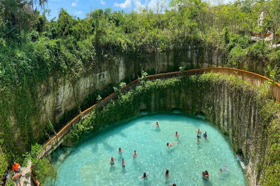 Cenote natural sinkhole pool surrounded by limestone cliffs, lush vegetation, and wooden railings with swimmers