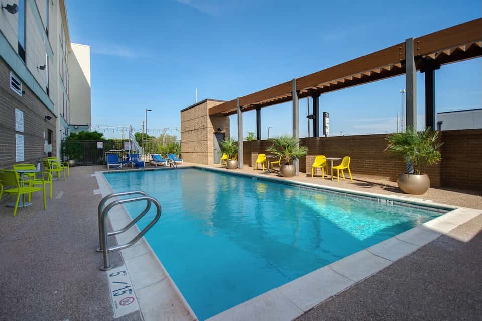 Resort pool with yellow loungers, brick building, pergola, and palm trees