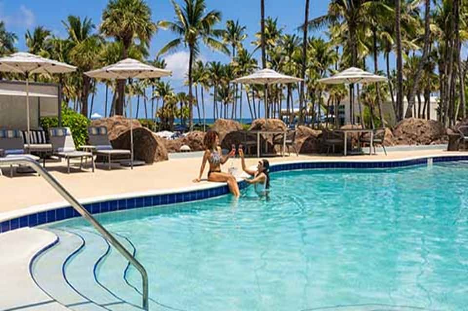 Beachfront resort pool with palm trees, loungers, umbrellas, and ocean backdrop