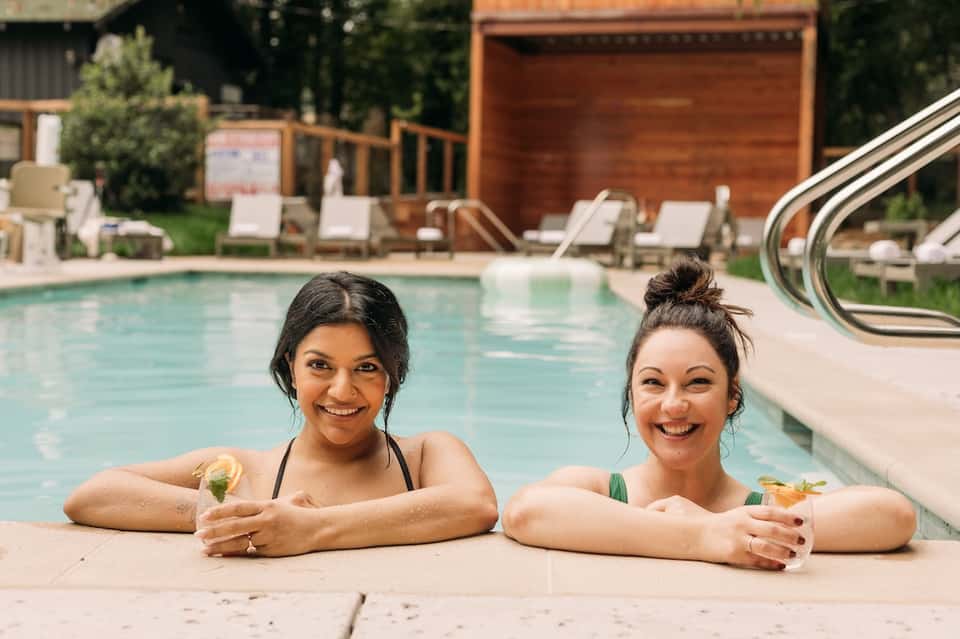 Two women in a resort pool holding tropical cocktails with loungers and landscaping visible