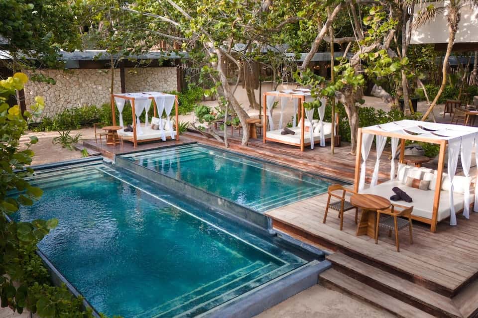 Luxe resort pool area with canopy beds, wooden deck, lush tropical landscaping and stone architecture