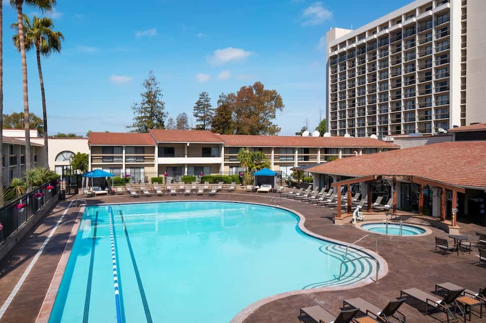 Large resort pool with lap lanes, jacuzzi, covered lounge area, and multi-story hotel building on sunny day