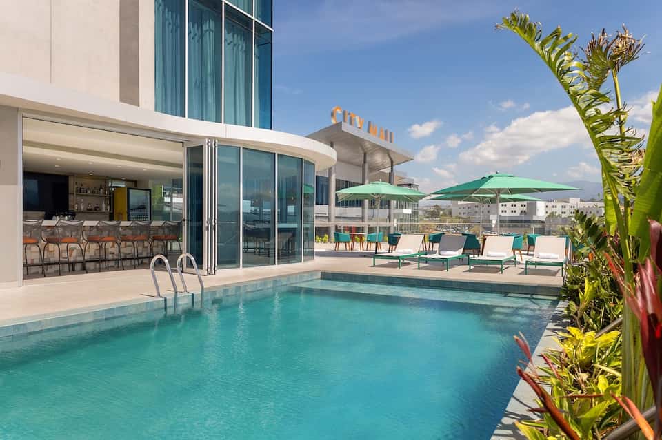Rooftop pool with modern bar, lounge chairs, green umbrellas, and city skyline views