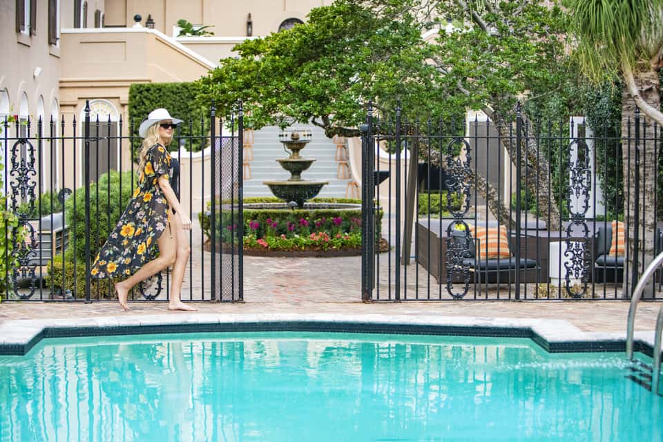 Woman in patterned dress near gated courtyard entrance with fountain, pool visible beyond