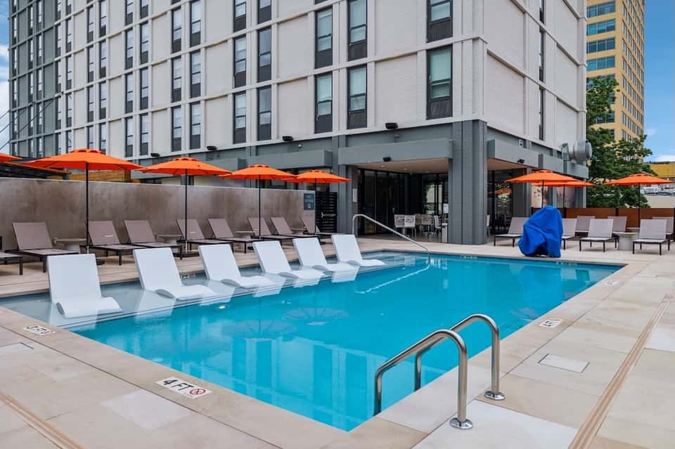 Urban hotel rooftop pool with white loungers, orange umbrellas, and city skyline backdrop
