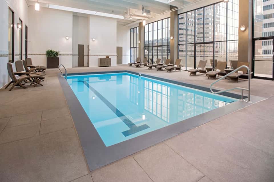 Modern indoor swimming pool with lap lanes, lounge chairs, and floor-to-ceiling windows with city views