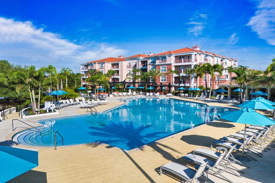 Large resort pool with pink building, blue umbrellas, lounge chairs, and palm trees