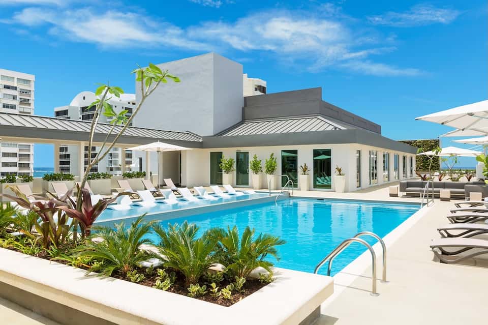 Resort pool and terrace with white umbrellas, tropical landscaping, and ocean view under blue sky