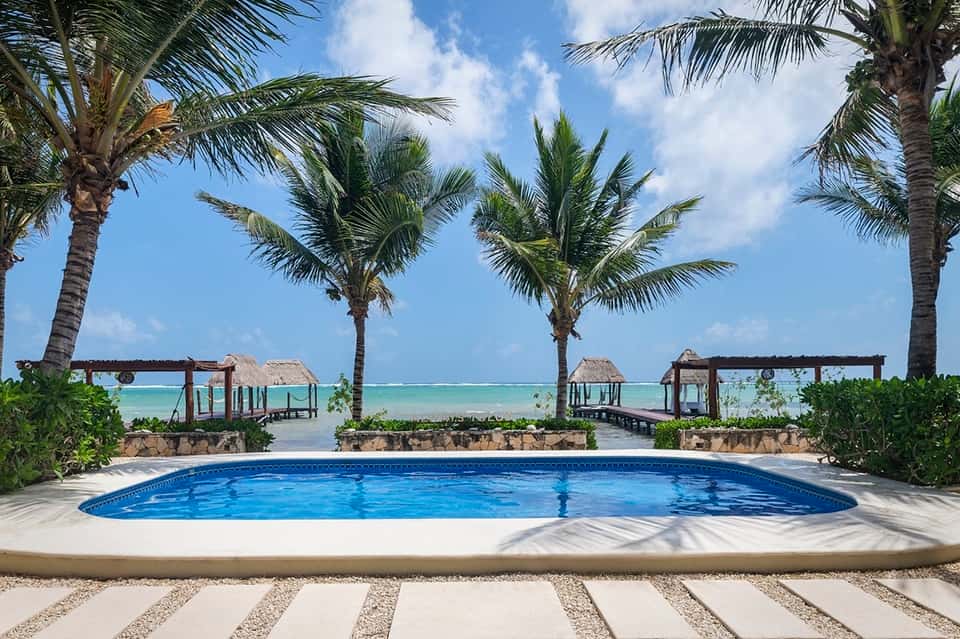 Beachfront resort pool with palm trees, ocean view, tiki bar structures, and tropical blue water
