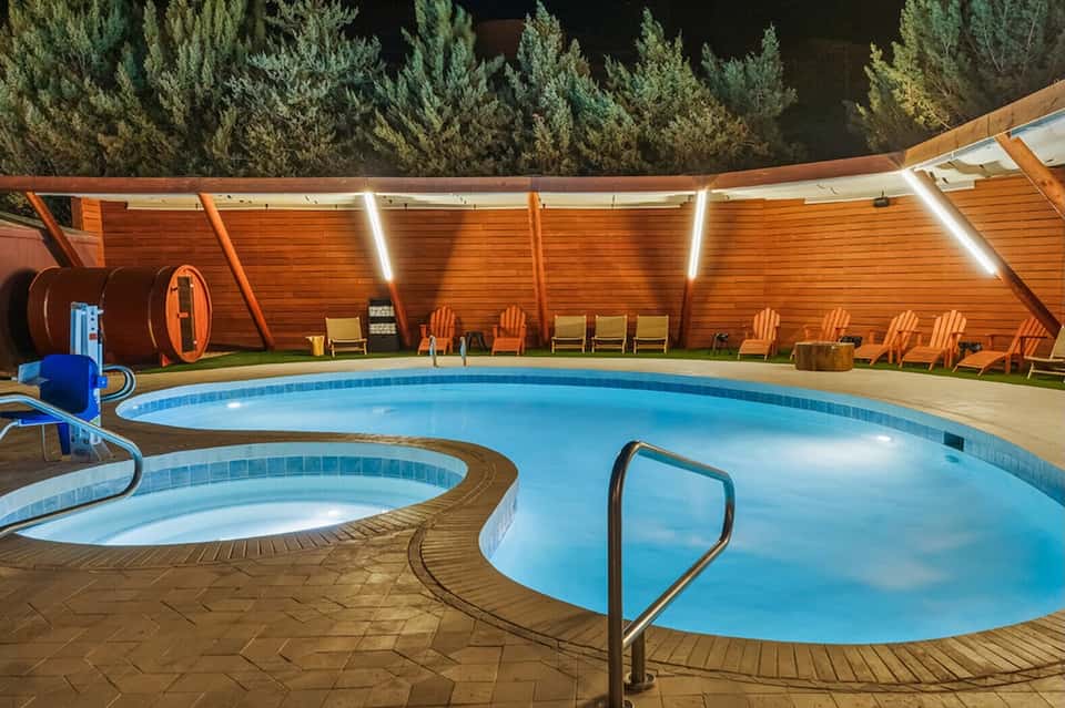 Indoor pool area with wooden walls, hot tub, orange Adirondack chairs, and pine forest backdrop