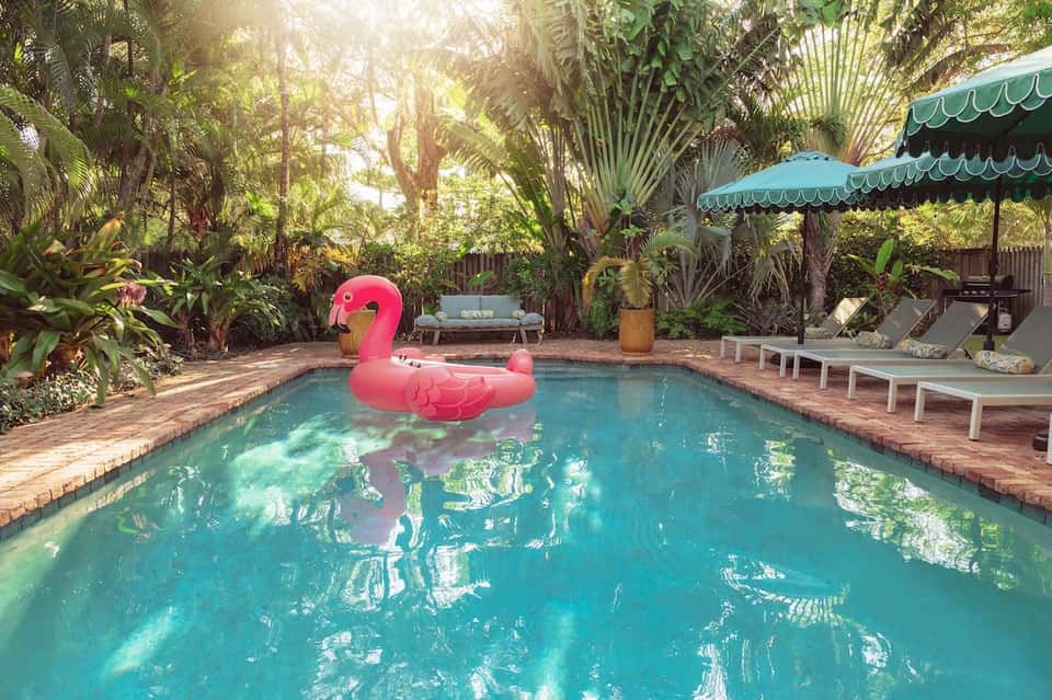 Tropical pool surrounded by palm trees, lounge chairs, turquoise umbrellas, and pink flamingo float