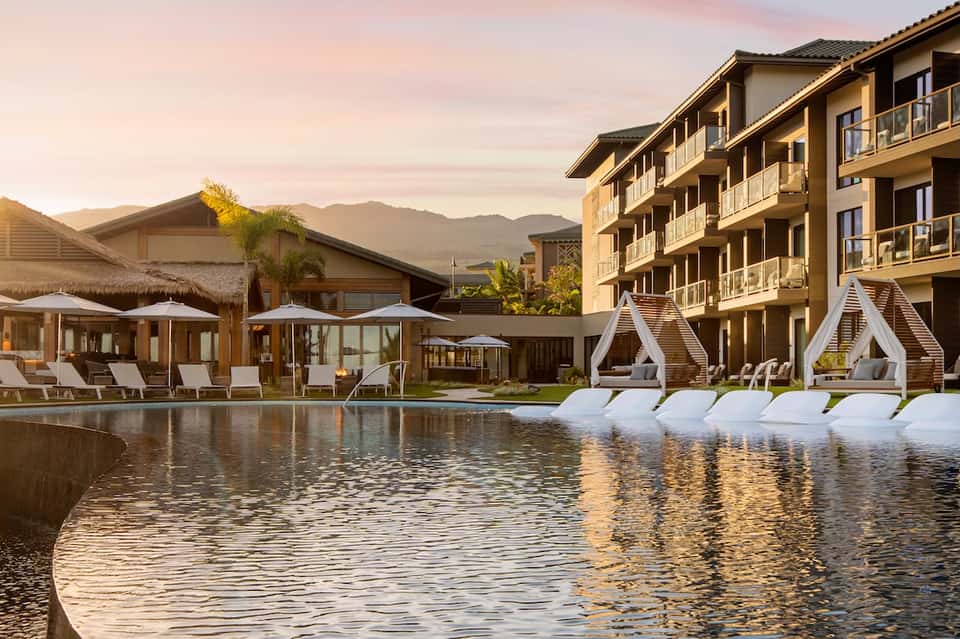 Luxury resort pool at sunset with mountain views, white cabanas, lounge chairs, and multi-story hotel building