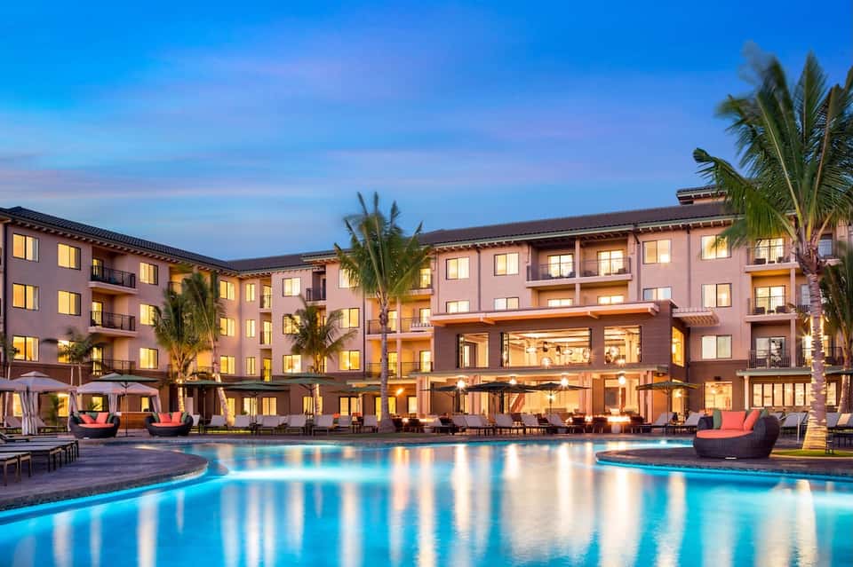 Luxury resort illuminated at dusk with glowing pool, pink building facade, palm trees, and lounge seating