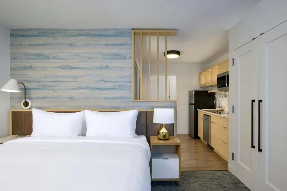 Compact hotel room with queen bed, kitchenette, wood accents, and blue wallpaper