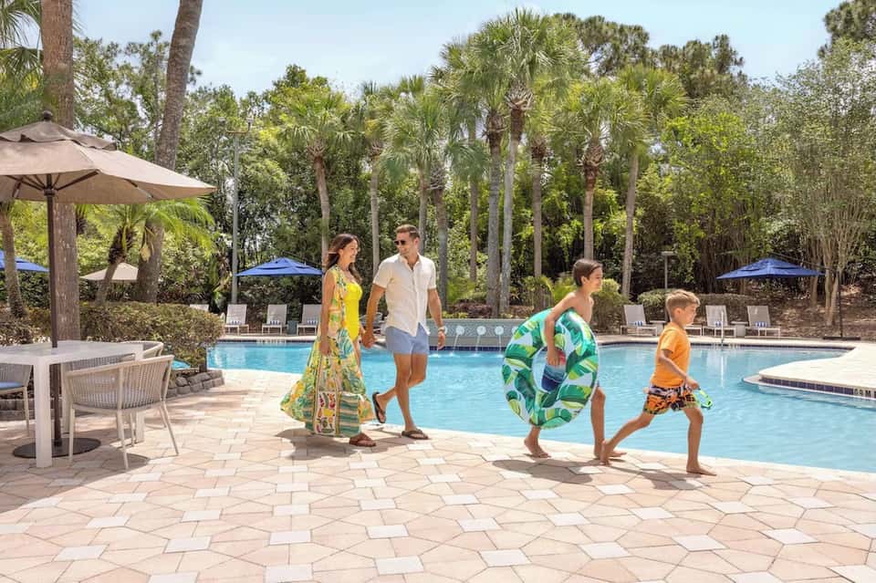 Resort pool area with family swimming, tropical trees, umbrellas, and lounge seating