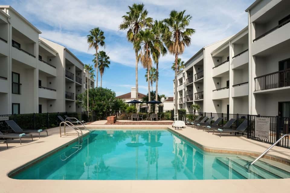 Resort pool with palm trees, multi-story white buildings, and lounge chairs