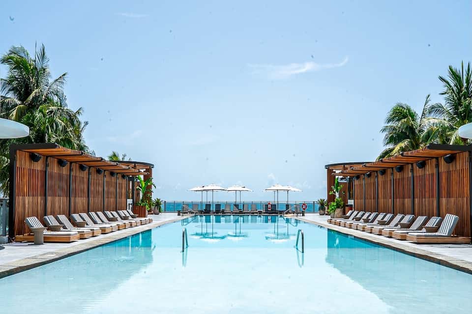 Luxury resort pool with wooden cabanas, striped loungers, palm trees, and ocean horizon view