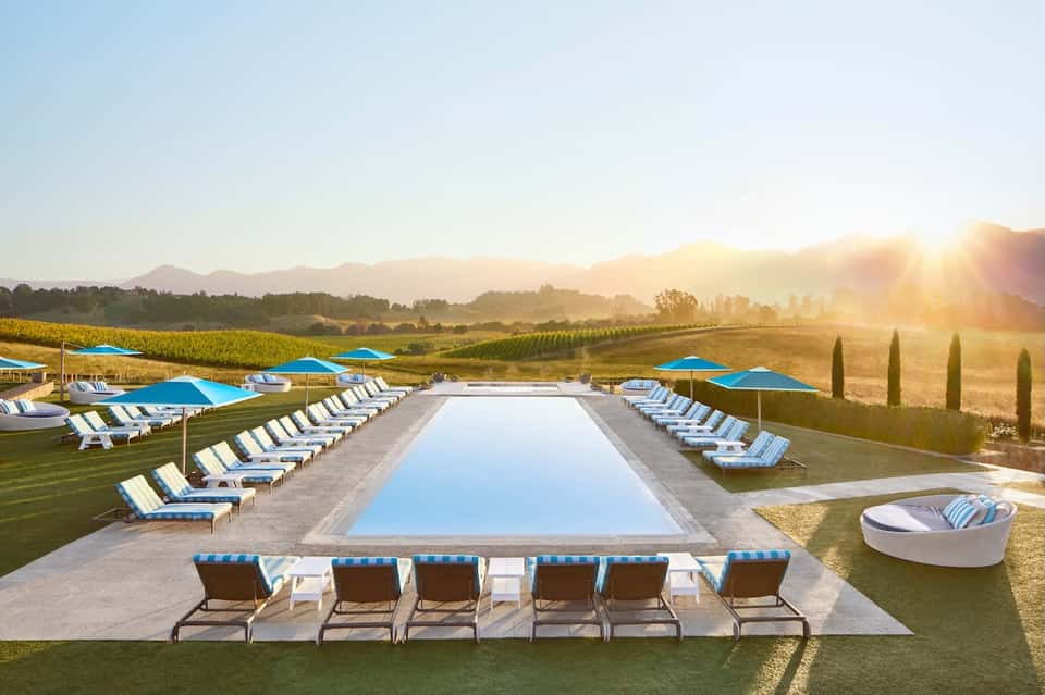 Resort lap pool with blue umbrellas and loungers overlooking vineyards and mountains at sunrise