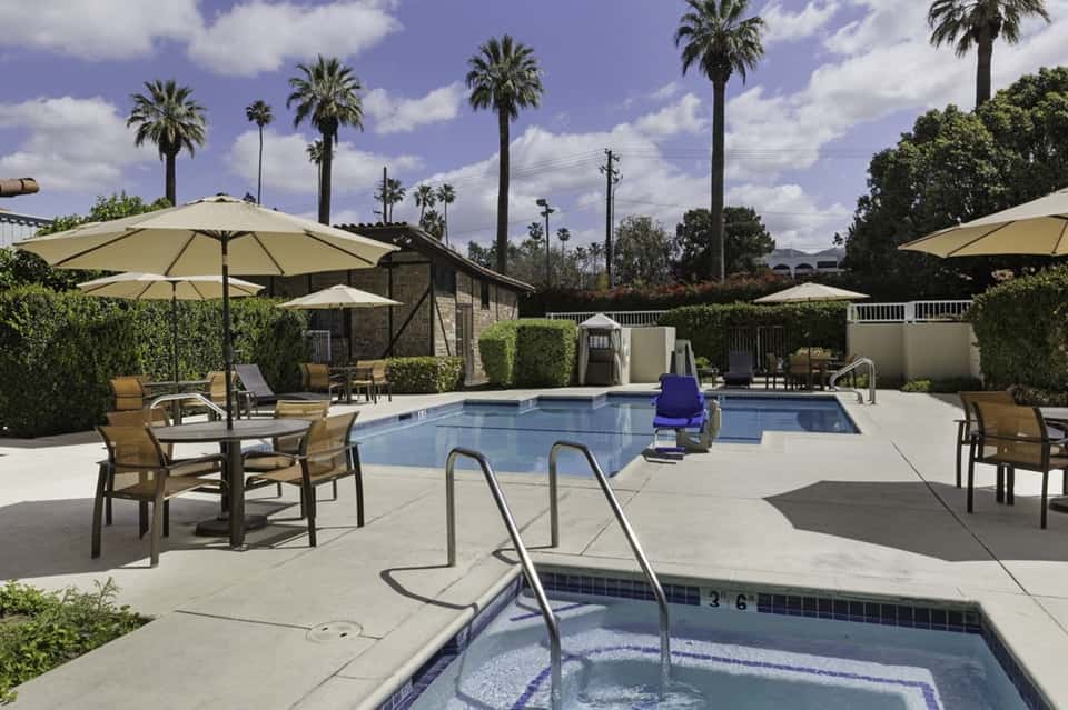 Resort pool area with beige umbrellas, palm trees, spa building, and hot tub in foreground