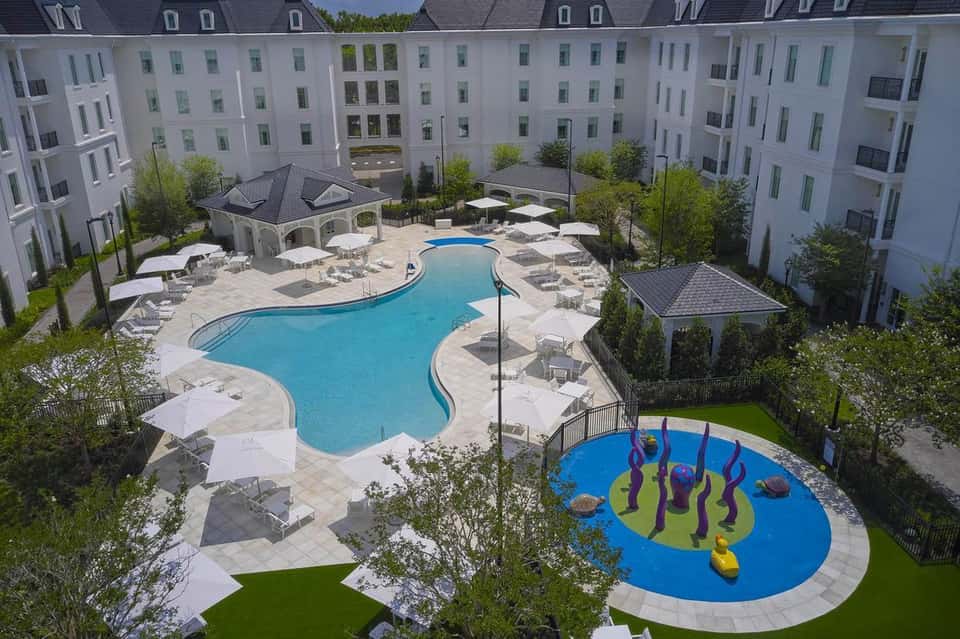 Aerial view of curved resort pool with splash pad, surrounding white buildings, and manicured landscaping