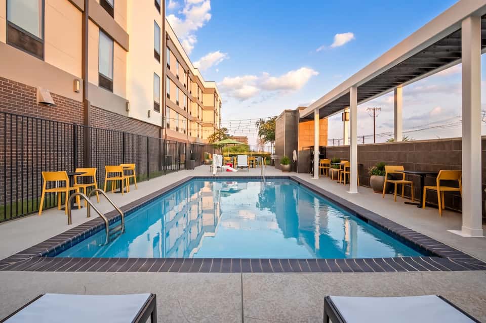 Modern narrow pool surrounded by yellow lounge chairs, white pergola, concrete deck, and contemporary hotel building