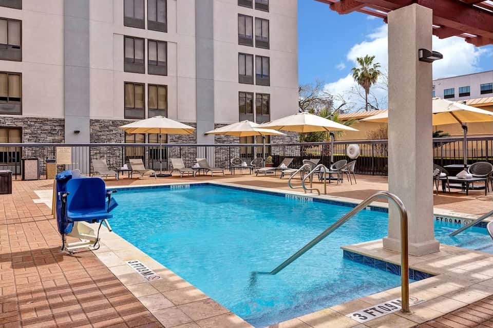 Hotel courtyard pool with blue water, tan umbrellas, lounge chairs, and multi-story building with stone accents.