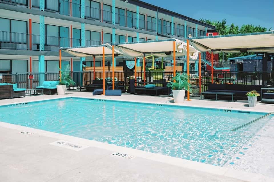 Outdoor pool with white shade sails, colorful umbrellas, and multi-story hotel building