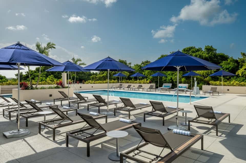 Upscale pool deck with blue umbrellas, multiple lounge chairs with cushions, and tropical landscaping