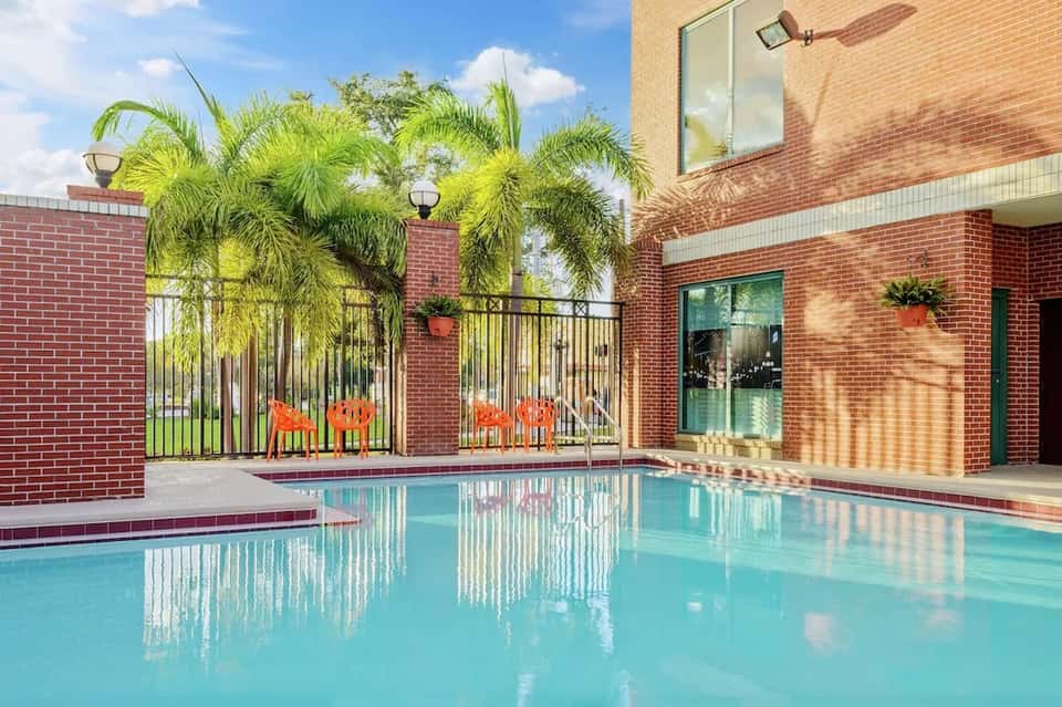 Residential pool with turquoise water, brick building, and palm trees beside glass doors