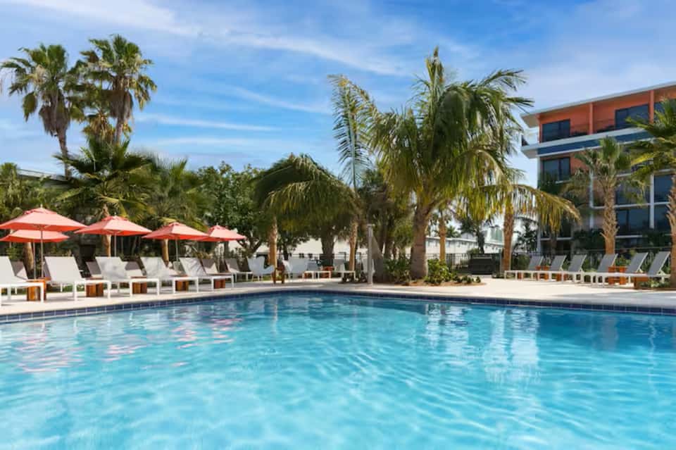 Resort pool surrounded by palm trees, red umbrellas, lounge chairs, and modern building