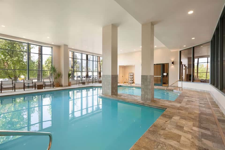 Indoor heated swimming pool with turquoise water, columns, large windows, and lounge seating area