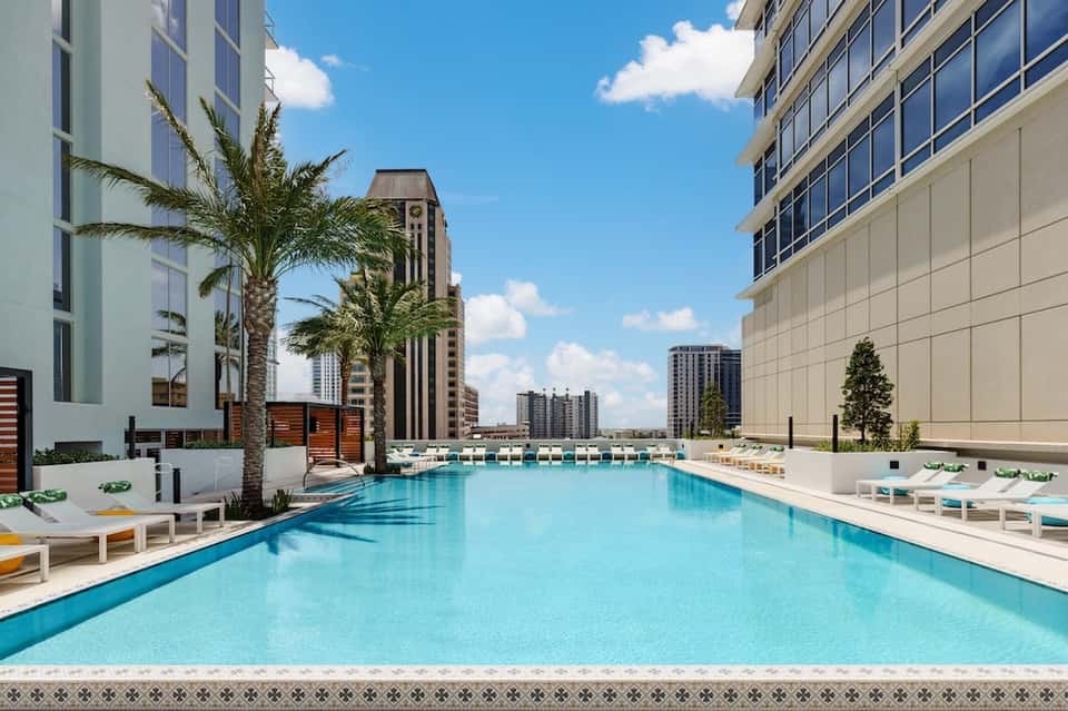 Urban resort pool with turquoise water, lounge chairs, palm trees, and modern high-rise buildings