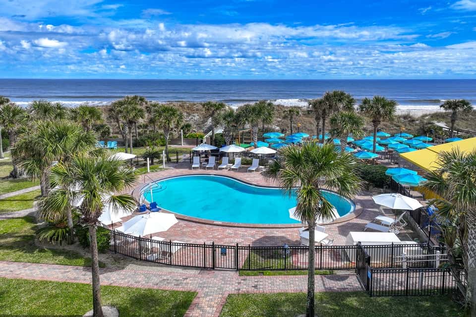 Beachfront resort pool with palm trees, blue umbrellas, ocean, and sandy beach view