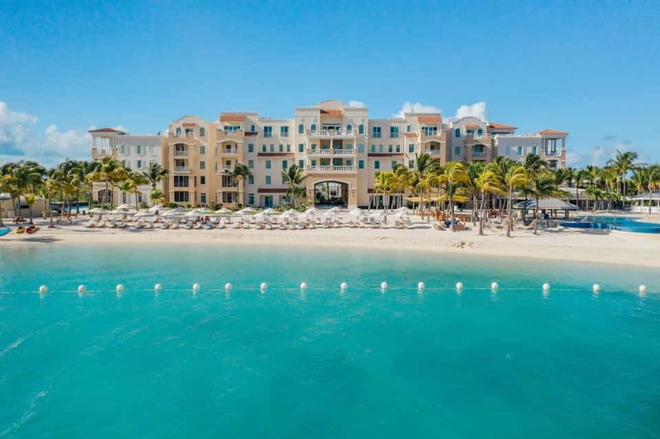 Colorful beachfront resort with multi-story pastel buildings, white sand beach, turquoise water, and palm trees