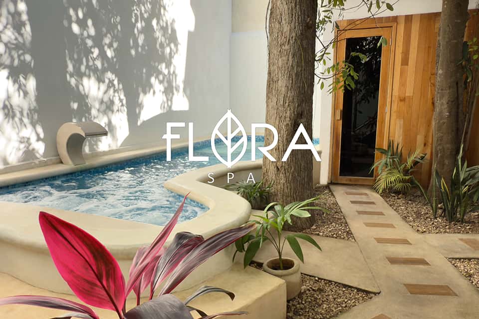 Flora Spa entrance with logo displayed on white wall, hot tub, wooden door, and tropical plants
