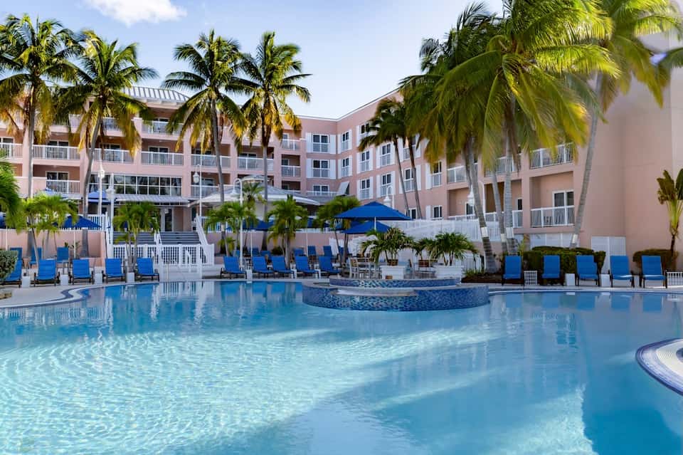 Luxury resort pool with turquoise water, blue umbrellas, loungers, and palm tree landscaping