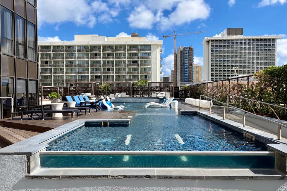 Rooftop pool with blue lounge chairs, city skyline, and modern high-rise buildings