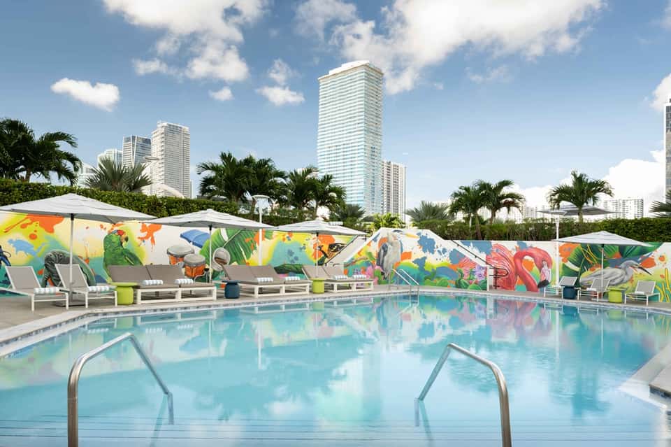 Colorful resort pool with vibrant mural backdrop, umbrellas, lounge chairs, and high-rise buildings in background